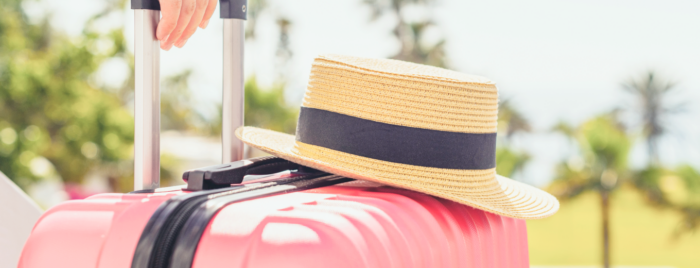 Close-up of a hat on a red suitcase held by a hand in front of sun-kissed palm trees